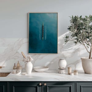 May include: A framed art print featuring a top-down view of a rowing team on a teal blue body of water. The artwork is displayed above a kitchen counter with a gold faucet, white marble backsplash, and a potted olive tree.