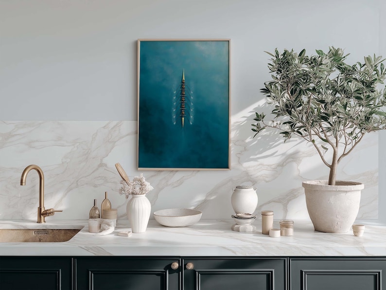 May include: A framed art print featuring a top-down view of a rowing team on a teal blue body of water. The artwork is displayed above a kitchen counter with a gold faucet, white marble backsplash, and a potted olive tree.
