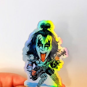 May include: A holographic sticker featuring a cartoon illustration of a musician with black hair, wearing black makeup and a black costume, playing a guitar. The musician has a long tongue sticking out. The sticker has a rainbow iridescent finish.