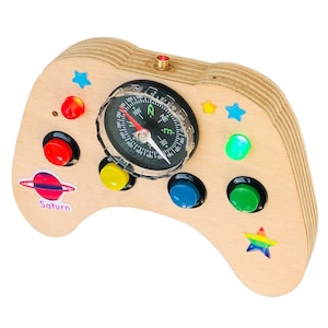 Op de afbeelding: Een houten speelcontroller met een kompas in het midden, vier kleurrijke knoppen en een afbeelding van de planeet Saturnus. De controller heeft een oplichtende ster en een oplichtende knop.