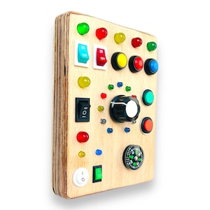 May include: Wooden sensory board with various interactive elements. Features toggle switches, push buttons, a rotary dial, and a compass. Includes colorful lights in red, green, and yellow. Designed for tactile exploration and learning.