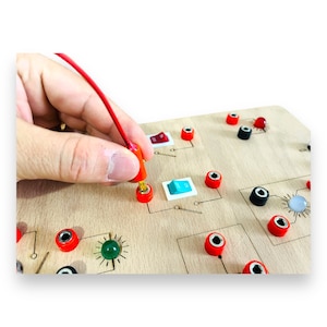 以下が含まれることがあります： 赤と黒の端子、スイッチ、緑色のライトなど、さまざまなコンポーネントを備えた木製の回路基板。赤いワイヤーが端子に接続されています。このボードは、おそらく電子工学について学ぶための教育目的で設計されています。