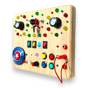 May include: Wooden activity board with interactive elements. Includes knobs, switches, buttons, and a spinning wheel with a star design. Features colourful, round, translucent accents. A red cord and keys are also present.