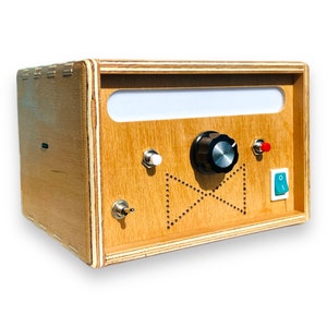 May include: A wooden box with a white rectangular opening, a black knob, a white button, a red button, and a green button. The box has a patterned design with a bow tie shape.