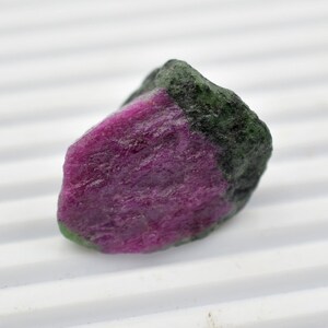 May include: A rough, uncut pink and green gemstone. The gemstone has a rough, natural texture and is a vibrant pink color with a green band across the top.