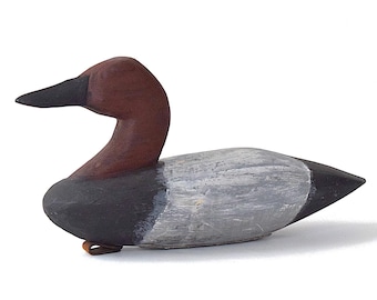 Puede incluir: Un señuelo de pato de madera tallado a mano. El pato tiene la cabeza y el cuello de color marrón oscuro, un pico negro y un cuerpo gris y negro. El señuelo está sobre una superficie blanca.