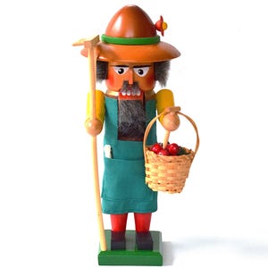 May include: Wooden nutcracker figurine of a farmer in a green apron and orange hat, holding a basket of red apples and a wooden tool. The figurine is painted with details like a gray beard and red boots. The base is green.