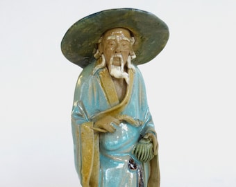 May include: A ceramic figurine of an elderly Asian man wearing a wide-brimmed hat and traditional robes. The figure is primarily turquoise with accents of brown and green. The man has a long white beard and is holding a small pouch.