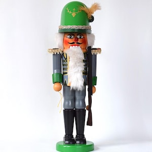 May include: A wooden nutcracker figurine in a hunter's uniform. The figurine has a green hat with a gold deer emblem and feather, a white beard, and a rifle. The base and hat are green, and the uniform is grey.