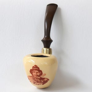 Puede incluir: Una pipa de tabaco vintage con un cuenco de color crema con una ilustración roja de un hombre con sombrero fumando en pipa. La pipa tiene una boquilla marrón oscuro y un accesorio de latón.