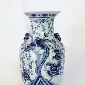 May include: A tall, blue and white porcelain vase with a flared rim and two side handles. The vase features a detailed design of a bird perched on a tree branch, surrounded by floral and botanical elements. The vase is approximately 14 inches tall.