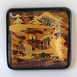 Puede incluir: Caja decorativa cuadrada con marco negro. La caja presenta una escena de paisaje detallada con una montaña, árboles, barcos y edificios en dorado sobre un fondo marrón y beige. La caja tiene un acabado brillante.