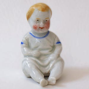 May include: A porcelain figurine of a seated baby with blonde hair, rosy cheeks, and blue accents on the collar and sleeves. The baby is wearing a white outfit. The figurine has a vintage aesthetic.
