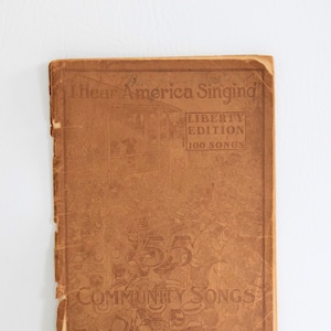 May include: A vintage book cover with the title "I Hear America Singing" and "Liberty Edition 100 Songs." The cover is a faded brown color with embossed text and illustrations. The book is titled "55 Community Songs" and published by C.C. Birchard & Co., Boston, Mass.
