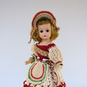 May include: A vintage doll wearing a red, white, and green crocheted dress with a matching hat. The doll has blonde hair and blue eyes.