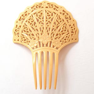 May include: A cream-colored hair comb with a fan-shaped top and long teeth. The top features an intricate floral and scrollwork design. The comb is made of a solid material and is set against a white background.