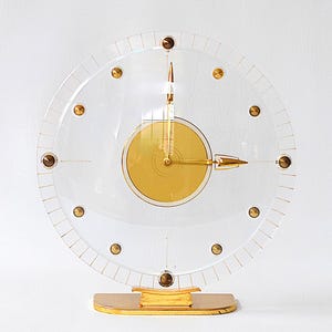 May include: A clear acrylic and gold-toned desk clock. The clock face is circular with gold accents and a gold center. The clock hands are gold. The base is rectangular and gold. The clock is a decorative item.