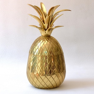 Puede incluir: Un recipiente decorativo dorado con forma de piña. El recipiente tiene un cuerpo texturizado con patrón de diamantes y una corona detallada con hojas. La piña es de metal y tiene un acabado brillante, que refleja la luz.