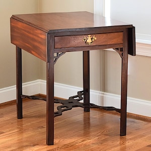 Puede incluir: Una mesa auxiliar de madera marrón oscuro con un solo cajón y una tapa abatible. La mesa tiene un tirador decorativo de latón en el cajón y soportes ornamentados. La mesa mide aproximadamente 61 cm de altura.