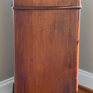 May include: A tall, antique wooden cabinet with a white top. The cabinet is made of dark brown wood with a rectangular shape. It has a decorative band around the middle and sits on small, rounded feet. The cabinet is positioned in a corner.