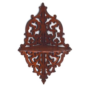 Può includere: Una mensola angolare in legno marrone scuro con intricati motivi a volute traforate. L'unità da parete presenta una superficie triangolare piatta per l'esposizione e un pannello posteriore decorativo a punta che si estende sopra e sotto.