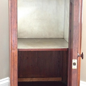May include: A vintage wooden cabinet with a drawer and a door. The cabinet is made of dark brown wood with a lighter interior. The door is open, revealing a shelf inside. The cabinet has a brass knob and a keyhole.