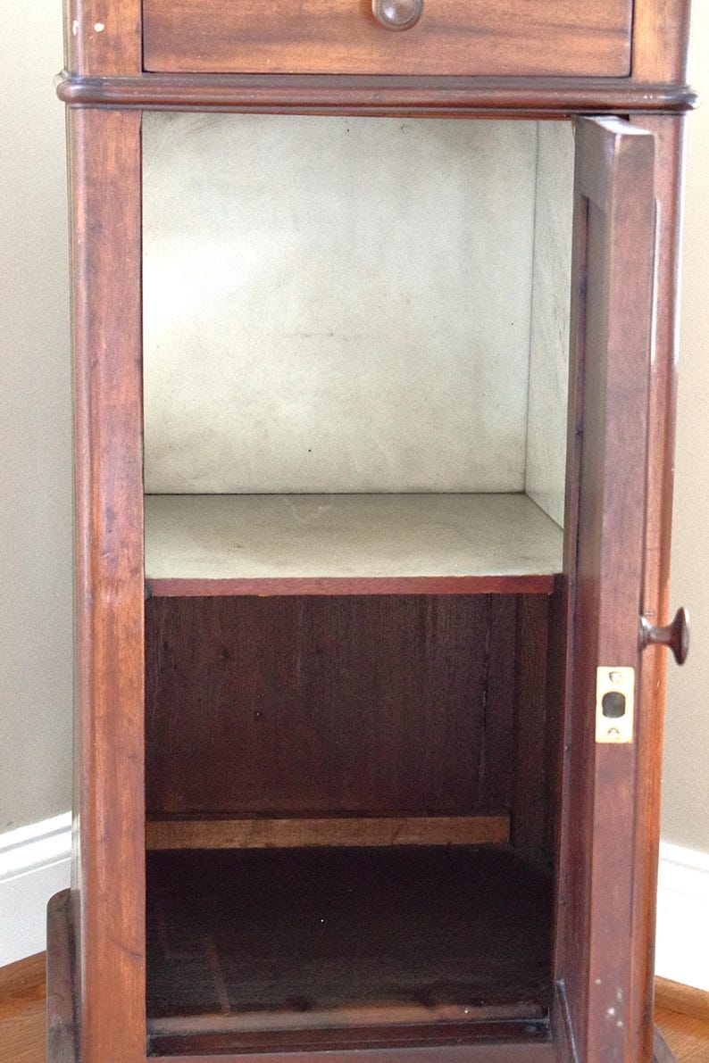 May include: A vintage wooden cabinet with a drawer and a door. The cabinet is made of dark brown wood with a lighter interior. The door is open, revealing a shelf inside. The cabinet has a brass knob and a keyhole.