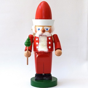 May include: A wooden Christmas nutcracker in the shape of a sailor, wearing a red uniform and hat, white beard, and holding a small green tree. The nutcracker stands on a green base.