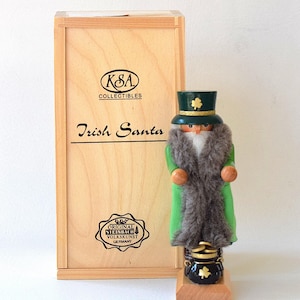 Peut inclure: Figurine en bois du Père Noël irlandais, vêtu d'un manteau vert avec une garniture en fourrure et d'un chapeau vert avec un trèfle doré. La figurine est sur un socle en bois avec un pot d'or noir. La boîte en bois indique "Irish Santa".
