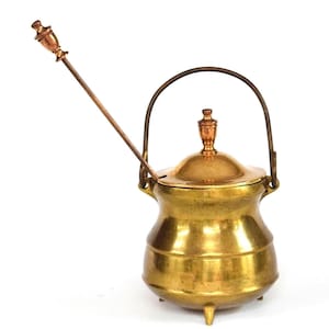Puede incluir: Una olla de latón vintage con tapa y asa, con una cuchara larga para remover. La olla tiene un cuerpo redondeado, tres patas pequeñas y un adorno decorativo en la tapa. El latón tiene un tono dorado cálido.