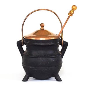 Puede incluir: Un pequeño caldero de hierro fundido negro con tapa y asa de cobre. El caldero tiene tres patas pequeñas y dos asas. Una cuchara para remover de color cobre y dorado descansa dentro.