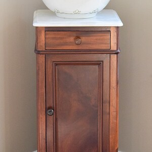 May include: A white porcelain pitcher and bowl set with gold trim, resting on a marble-topped wooden cabinet. The cabinet is dark brown with a drawer and a door. The set is likely for washing or decorative purposes.