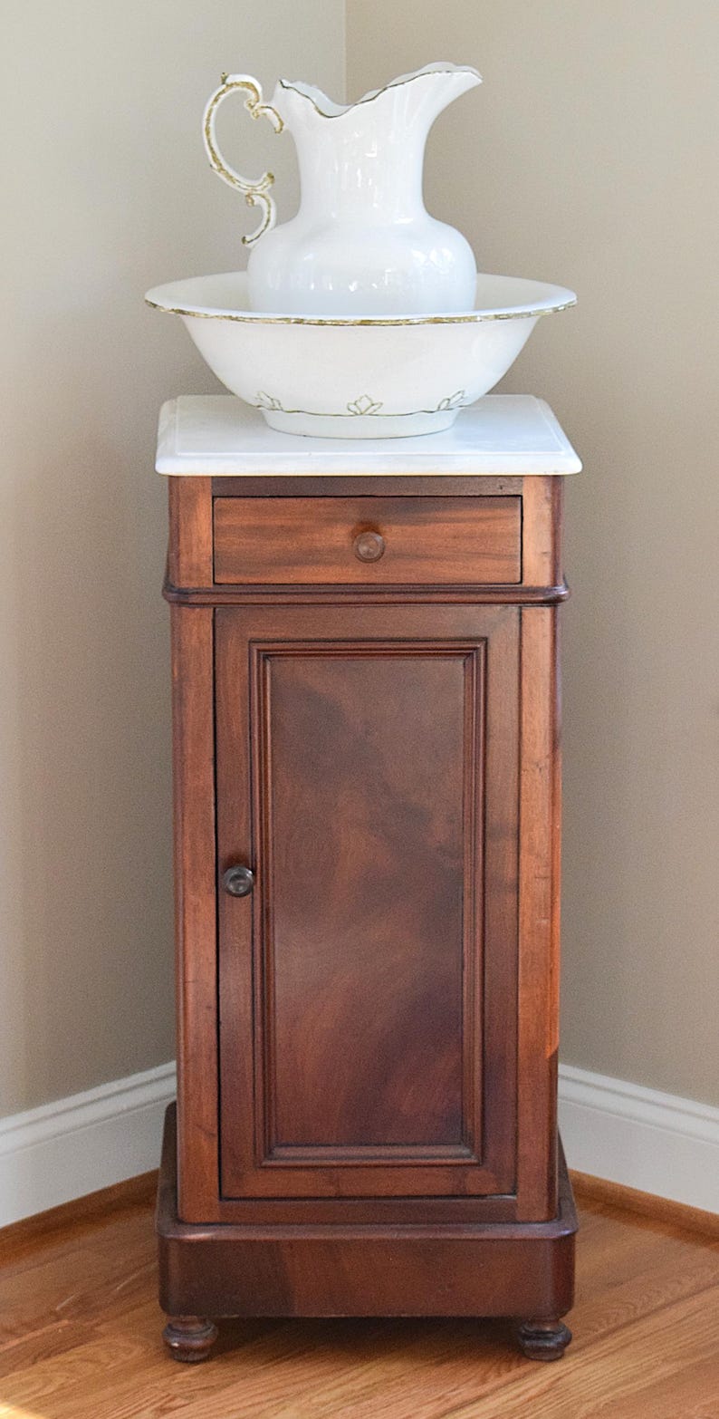 May include: A white porcelain pitcher and bowl set with gold trim, resting on a marble-topped wooden cabinet. The cabinet is dark brown with a drawer and a door. The set is likely for washing or decorative purposes.
