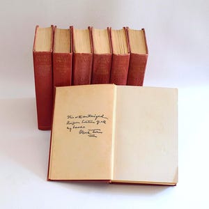 Könnte beinhalten: Eine Sammlung antiker Bücher mit roten Einbänden, darunter Titel von Mark Twain. Ein Buch ist geöffnet und zeigt handschriftlichen Text und eine Unterschrift. Die Bücher sind auf einer weißen Oberfläche angeordnet und zeigen ihr gealtertes Aussehen.