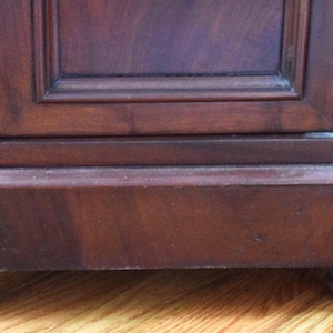 May include: Close-up of a dark brown wooden cabinet base with a rectangular design. The cabinet has a decorative trim and sits on four rounded feet. The wood grain is visible, and the finish appears to be glossy.