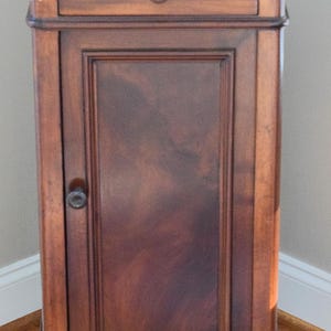 May include: A tall, antique wooden cabinet with a white marble top. The cabinet has a single drawer and a door with a dark, reddish-brown finish. The door has a small, round handle. The cabinet is supported by short, rounded feet.