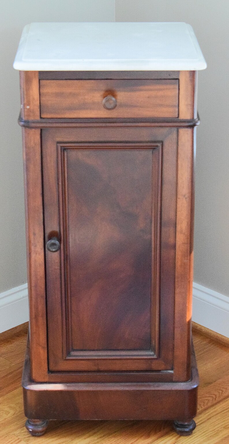 May include: A tall, antique wooden cabinet with a white marble top. The cabinet has a single drawer and a door with a dark, reddish-brown finish. The door has a small, round handle. The cabinet is supported by short, rounded feet.