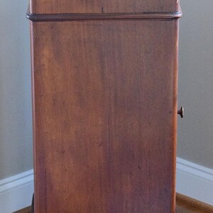 May include: A tall, antique wooden cabinet with a white top. The cabinet has a rich, dark brown finish and a small, round handle. The cabinet is rectangular and stands on four small, rounded feet. The wood grain is visible.