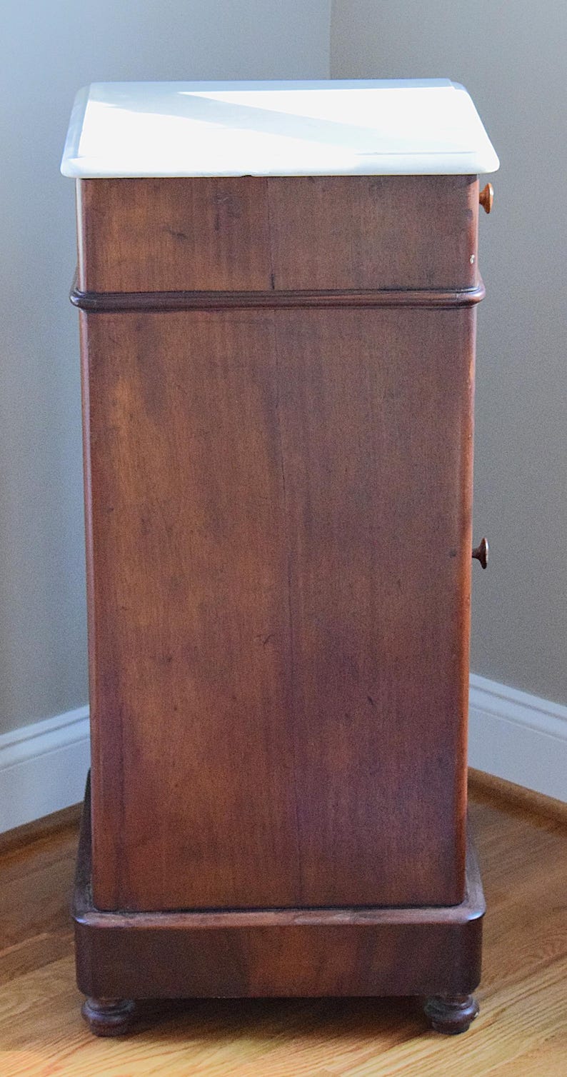 May include: A tall, antique wooden cabinet with a white top. The cabinet has a rich, dark brown finish and a small, round handle. The cabinet is rectangular and stands on four small, rounded feet. The wood grain is visible.