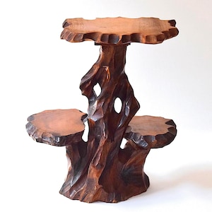 May include: A dark brown wooden display stand featuring three tiered platforms with uneven, carved edges. The central support structure is shaped like a gnarled tree trunk with a naturalistic, textured surface and a visible hollow opening in the center.