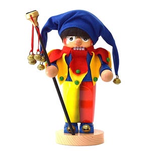 May include: A wooden jester figurine with a blue hat, red and yellow outfit, and a staff with bells. The figurine has a painted face with a wide smile and is standing on a wooden base. The jester is a decorative item.