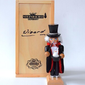 May include: A wooden Wizard nutcracker with a black top hat, red-trimmed black coat, and a white rabbit in a hat. The nutcracker stands on a wooden base next to a wooden box with the word "Wizard" and "Steinbach Germany" printed on it.