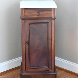May include: A tall, antique wooden cabinet with a white top. The cabinet has a single drawer with a round knob, a door with a dark wood grain, and sits on small, rounded feet. The cabinet is in a corner of a room with hardwood floors.