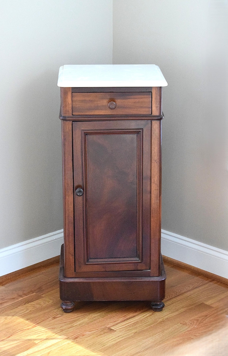 May include: A tall, antique wooden cabinet with a white top. The cabinet has a single drawer with a round knob, a door with a dark wood grain, and sits on small, rounded feet. The cabinet is in a corner of a room with hardwood floors.