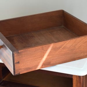 May include: A wooden drawer with a warm brown finish, featuring a small, round handle. The drawer is open, revealing its interior. The dovetail joints are visible on the corners. The drawer is resting on a white surface.