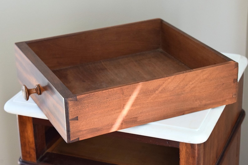 May include: A wooden drawer with a warm brown finish, featuring a small, round handle. The drawer is open, revealing its interior. The dovetail joints are visible on the corners. The drawer is resting on a white surface.