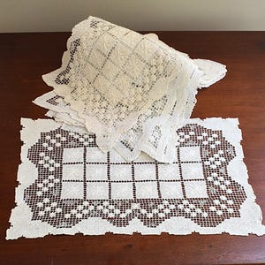 Puede incluir: Una pila de tapetes de encaje rectangulares de color blanco roto con un patrón central de cuadrícula de cuadrados sólidos rodeado por un intrincado borde calado con bordes festoneados. Un tapete está extendido en primer plano mostrando el detalle.