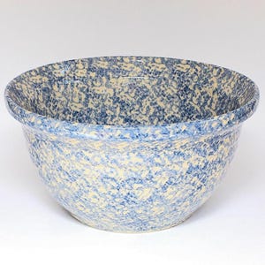 May include: A round ceramic mixing bowl featuring a speckled pattern with a light cream base and mottled blue paint throughout. The bowl has a wide, open top with a thick, rounded rim and a deep, curved basin, showcasing a consistent finish.