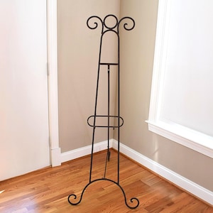 May include: A tall, dark brown metal display easel featuring a slender, tripod-style frame with curved scrollwork accents at the top and base. The structure includes a central adjustable support bar and a horizontal shelf for holding items securely.