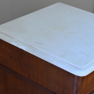 May include: A close-up of a wooden cabinet with a white marble top. The cabinet is made of dark brown wood with a curved design. The marble top has a slightly rounded edge.